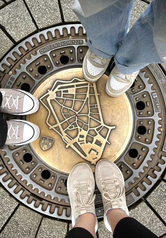 Füße stehen rund um den Innenstadtplan auf einem Gullideckel in der Oldenburger Innenstadt. 