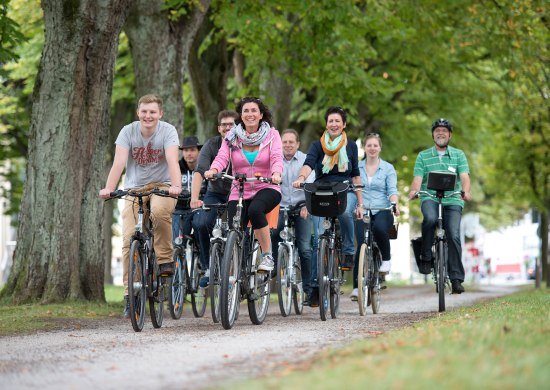 Eine Gruppe Radfahrer im Rahmen einer Stadtführung mit dem Fahrrad auf einem baumumsäumten Weg in Oldenburg unterwegs. 