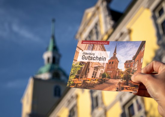 Eine Hand hält die Klappkarte des Oldenburg-Gutscheins vor dem Oldenburger Schloss