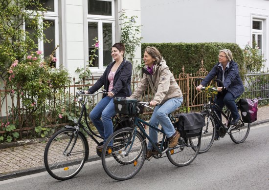 Drei Fahrradfahrerinnen in der Stadt. Im Hintergrund sind die typischen Oldenburger Hundehütten.