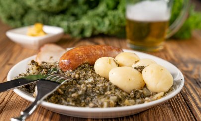 Traditionelles Tellergericht mit Grünkohl, Pinkelwurst und Kartoffeln. Dazu eine Schale Senf und ein Bier auf einem Holztisch.