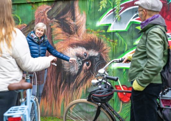 Gästeführerin Martina Petzalis vor einem Graffiti. Im Vordergrund zwei Teilnehmende der Tour mit ihrem Fahrrad. 