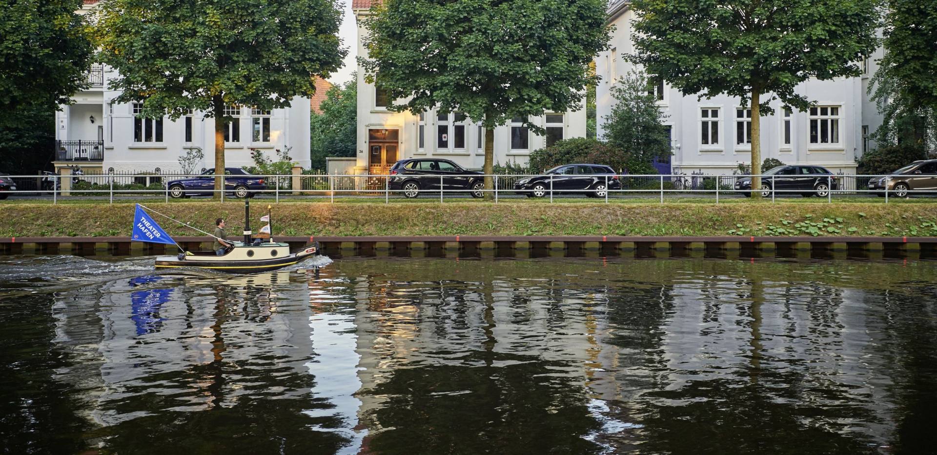 Ein kleines Boot fährt auf dem Küstenkanal durch Oldenburg vor drei Bäumen und drei Häusern.