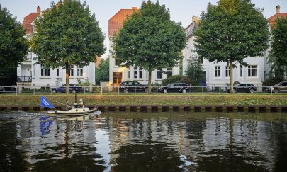 Ein kleines Boot fährt auf dem Küstenkanal durch Oldenburg vor drei Bäumen und drei Häusern.