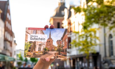 Eine Hand hält den Oldenburg Gutschein. Im Hintergrund die Oldenburger Fußgängerzone. Eine Hand hält den Oldenburg Gutschein. Im Hintergrund die Oldenburger Fußgängerzone.