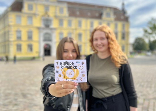 Mariia Strateichuk und Merle Stührenberg halten die Postkarte zur Aktion "Kultursnacks" vor dem Landesmuseum Kunst & Kultur in die Kamera. 